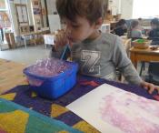 Child blowing bubbles into purple liquid and using to paint