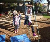 Child climbing a rope ladder outside, with children lining up behind her to go next